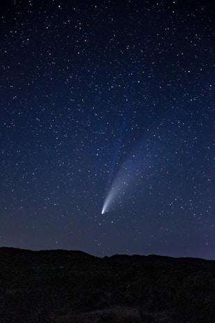 A clear night sky filled with stars, featuring a bright comet with a vivid tail extending across the sky. The silhouette of a landscape with gentle hills is visible at the bottom, emphasizing the vastness of the sky.