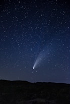 A clear night sky filled with stars, featuring a bright comet with a vivid tail extending across the sky. The silhouette of a landscape with gentle hills is visible at the bottom, emphasizing the vastness of the sky.