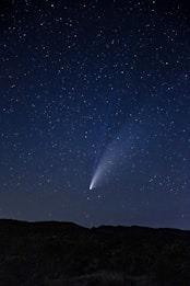 A clear night sky filled with stars, featuring a bright comet with a vivid tail extending across the sky. The silhouette of a landscape with gentle hills is visible at the bottom, emphasizing the vastness of the sky.