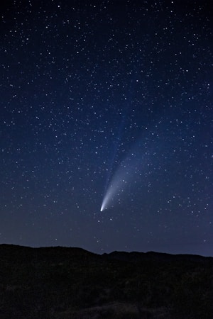A clear night sky filled with stars, featuring a bright comet with a vivid tail extending across the sky. The silhouette of a landscape with gentle hills is visible at the bottom, emphasizing the vastness of the sky.
