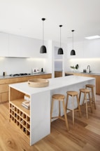 A modern kitchen with a minimalist design featuring a white island in the center accompanied by four wooden bar stools. The kitchen includes a countertop with a gas stove, sleek cabinetry, and three pendant lights hanging above the island. The floor is wooden, and a few decorative elements like a plant and bottles are placed on the counter. An open shelf with a built-in wine rack is part of the island.
