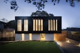 A modern two-story house with a sleek, minimalist design. The upper level features large vertical windows and a dark facade, while the lower level has a lighter appearance with smaller windows. The surrounding area includes a well-manicured lawn and a paved driveway, with ambient lighting creating a warm glow in the evening setting.