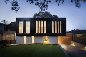 A modern two-story house with a sleek, minimalist design. The upper level features large vertical windows and a dark facade, while the lower level has a lighter appearance with smaller windows. The surrounding area includes a well-manicured lawn and a paved driveway, with ambient lighting creating a warm glow in the evening setting.