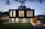 A modern two-story house with a sleek, minimalist design. The upper level features large vertical windows and a dark facade, while the lower level has a lighter appearance with smaller windows. The surrounding area includes a well-manicured lawn and a paved driveway, with ambient lighting creating a warm glow in the evening setting.