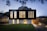 A modern two-story house with a sleek, minimalist design. The upper level features large vertical windows and a dark facade, while the lower level has a lighter appearance with smaller windows. The surrounding area includes a well-manicured lawn and a paved driveway, with ambient lighting creating a warm glow in the evening setting.