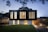 A modern two-story house with a sleek, minimalist design. The upper level features large vertical windows and a dark facade, while the lower level has a lighter appearance with smaller windows. The surrounding area includes a well-manicured lawn and a paved driveway, with ambient lighting creating a warm glow in the evening setting.