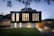 A modern two-story house with a sleek, minimalist design. The upper level features large vertical windows and a dark facade, while the lower level has a lighter appearance with smaller windows. The surrounding area includes a well-manicured lawn and a paved driveway, with ambient lighting creating a warm glow in the evening setting.