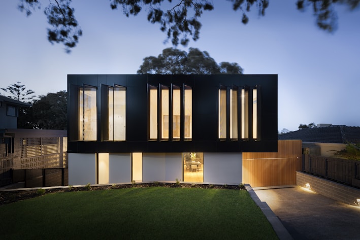 A modern two-story house with a sleek, minimalist design. The upper level features large vertical windows and a dark facade, while the lower level has a lighter appearance with smaller windows. The surrounding area includes a well-manicured lawn and a paved driveway, with ambient lighting creating a warm glow in the evening setting.