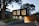 A modern two-story house with a sleek, black upper facade and a wooden lower section. Large vertical windows allow warm light to shine through, creating a welcoming atmosphere. The property is surrounded by trees and has a neatly maintained lawn and stone pathway leading to the entrance.