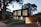 A modern two-story house with a sleek, black upper facade and a wooden lower section. Large vertical windows allow warm light to shine through, creating a welcoming atmosphere. The property is surrounded by trees and has a neatly maintained lawn and stone pathway leading to the entrance.