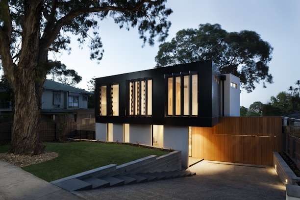 A modern two-story house with a sleek, black upper facade and a wooden lower section. Large vertical windows allow warm light to shine through, creating a welcoming atmosphere. The property is surrounded by trees and has a neatly maintained lawn and stone pathway leading to the entrance.