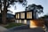 A modern two-story house with a sleek, black upper facade and a wooden lower section. Large vertical windows allow warm light to shine through, creating a welcoming atmosphere. The property is surrounded by trees and has a neatly maintained lawn and stone pathway leading to the entrance.