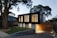 A modern two-story house with a sleek, black upper facade and a wooden lower section. Large vertical windows allow warm light to shine through, creating a welcoming atmosphere. The property is surrounded by trees and has a neatly maintained lawn and stone pathway leading to the entrance.