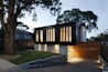 A modern two-story house with a sleek, black upper facade and a wooden lower section. Large vertical windows allow warm light to shine through, creating a welcoming atmosphere. The property is surrounded by trees and has a neatly maintained lawn and stone pathway leading to the entrance.