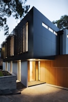 A modern, minimalist two-story house with a combination of dark panels and wooden slats on the exterior. It features large windows and a well-lit entrance. There are trees around the building, adding a touch of nature to the contemporary design.