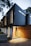 A modern, minimalist two-story house with a combination of dark panels and wooden slats on the exterior. It features large windows and a well-lit entrance. There are trees around the building, adding a touch of nature to the contemporary design.