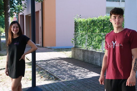 Two young adults stand outdoors on a sunny day, surrounded by greenery and buildings. The woman is wearing a black dress and smiling, while the man is casually dressed in a burgundy t-shirt and black shorts.
