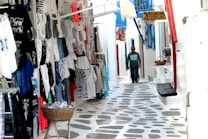 A narrow cobblestone alleyway lined with various shops, displaying colorful clothing and souvenirs on racks outside. To the right, there's a wooden bench and a small basket with items. A person wearing sunglasses and a green shirt walks down the lane. Brightly painted doors and shutters in white and blue give the scene a Mediterranean feel.
