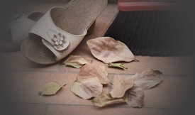 A pair of beige sandals adorned with a floral decoration rest on a tiled floor, surrounded by scattered dried leaves. A broom with dark bristles is placed nearby, suggesting the setting of an outdoor patio or porch in need of cleaning.