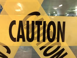 Yellow caution tape with bold black letters spelling 'CAUTION' is prominently displayed. The tape is positioned against a background that appears to be a parking garage or similar enclosed area, faintly visible through the tape.