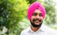 A smiling man with a beard wearing a bright pink turban and a white shirt. The background is a lush green, likely a garden or park, which is blurred to emphasize the focus on the man.