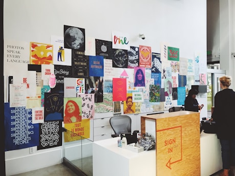 A wall adorned with a diverse collection of colorful posters and artwork featuring various inspirational quotes and imagery. In the foreground, there is a wooden sign-in desk with people interacting nearby.