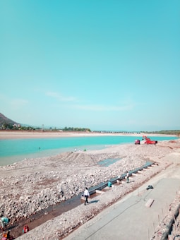 A clear turquoise river flows alongside a rocky embankment under a vast blue sky. Several people are engaged in construction activities near the riverbank, with a red truck and some construction materials visible. The background features green trees and a distant bridge crossing the water.