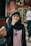 A person wearing a hijab and cap is eating a snack, standing in front of a store with a sign advertising a 40% discount on shoes. Another person is in the background, walking by and holding a phone.