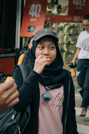 A person wearing a hijab and cap is eating a snack, standing in front of a store with a sign advertising a 40% discount on shoes. Another person is in the background, walking by and holding a phone.
