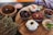 A rustic wooden board holds an assortment of small dishes filled with various dips, surrounded by natural herbs, a bowl of sea salt, and a wooden bowl of liquid. The dishes contain items like olives, sun-dried tomatoes, creamy white dip with a chili pepper, a beet-based dip with a walnut, and a basil leaf garnishing another dish. A checkered cloth is partially visible in the background.