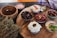 A rustic wooden board holds an assortment of small dishes filled with various dips, surrounded by natural herbs, a bowl of sea salt, and a wooden bowl of liquid. The dishes contain items like olives, sun-dried tomatoes, creamy white dip with a chili pepper, a beet-based dip with a walnut, and a basil leaf garnishing another dish. A checkered cloth is partially visible in the background.