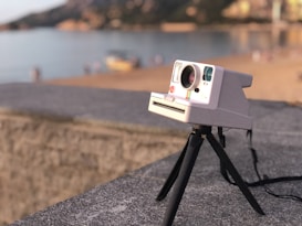 A vintage instant camera is mounted on a tripod overlooking a peaceful beach scene. In the background, the coastal landscape and calm waters are slightly blurred, suggesting a serene and nostalgic atmosphere.