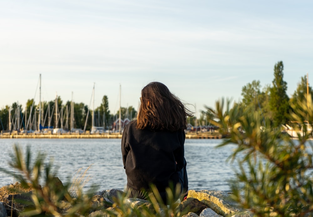 woman in black jacket sitting on rock near body of water during daytime