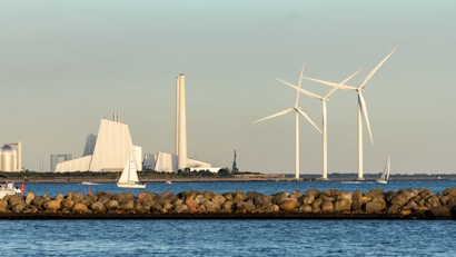 A coastal scene featuring a rocky barrier, several white sailboats on the water, and wind turbines in the background. A large industrial structure is also visible along the shoreline.