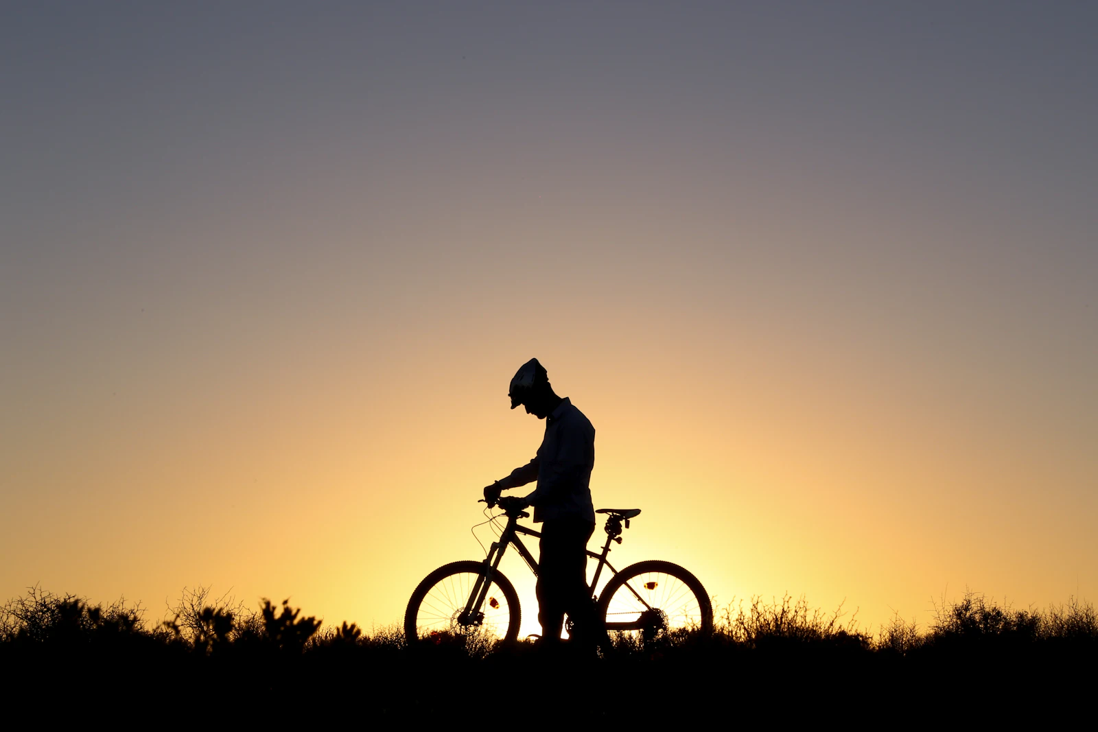 Silhouette of a rider standing beside a mountain bike on a ridge at sunset
