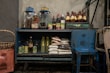 A rustic blue cabinet filled with various groceries and kitchen supplies. On top, there are two large glass jars and several bottles filled with sauces and beverages. Inside the cabinet, there are bags of rice, bottles of cooking oil, canned goods, and other food items. Beside the cabinet is a large woven basket, and a small blue stool is placed nearby.