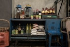 A rustic blue cabinet filled with various groceries and kitchen supplies. On top, there are two large glass jars and several bottles filled with sauces and beverages. Inside the cabinet, there are bags of rice, bottles of cooking oil, canned goods, and other food items. Beside the cabinet is a large woven basket, and a small blue stool is placed nearby.