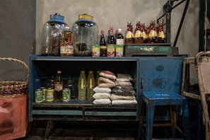 A rustic blue cabinet filled with various groceries and kitchen supplies. On top, there are two large glass jars and several bottles filled with sauces and beverages. Inside the cabinet, there are bags of rice, bottles of cooking oil, canned goods, and other food items. Beside the cabinet is a large woven basket, and a small blue stool is placed nearby.