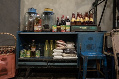 A rustic blue cabinet filled with various groceries and kitchen supplies. On top, there are two large glass jars and several bottles filled with sauces and beverages. Inside the cabinet, there are bags of rice, bottles of cooking oil, canned goods, and other food items. Beside the cabinet is a large woven basket, and a small blue stool is placed nearby.