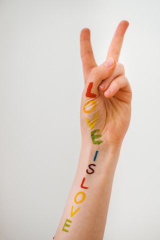 A hand is raised with fingers making a peace sign. The forearm has the phrase 'LOVE IS LOVE' written in colorful letters, each letter in a different color of the rainbow.