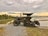 An agricultural tractor with caterpillar tracks is parked on a gravel path beside a rice field. The field is filled with water, reflecting the cloudy evening sky. Palm trees line the horizon, adding a tropical atmosphere to the rural setting.