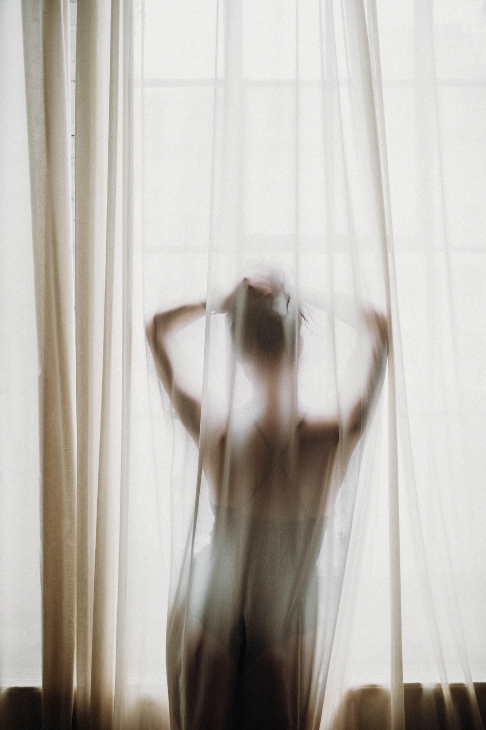 Fine-art silhouette of a figure behind a sheer curtain backlit by soft window light.