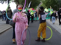 A person in a pink jumpsuit and rainbow face mask skillfully spins a hula hoop. Another person, dressed in colorful attire with a hat and 'Carbon Neutral Workforce' vest, stands nearby with hula hoops. Behind them, a group of people, some wearing masks and holding banners and flags, are gathered on the street, suggesting a protest or rally atmosphere. Trees line the street, and a few police officers are visible in the background.