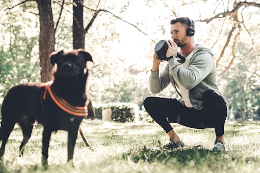 A man is squatting outdoors holding a kettlebell, wearing headphones and a gray hoodie. A black dog with an orange harness is in the foreground. Sunlight filters through the trees, creating a serene and active outdoor setting.