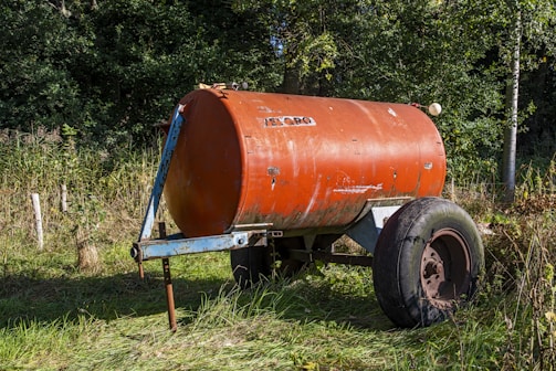 A rusty, orange cylindrical tank with wheels is placed on grassy terrain, surrounded by dense green foliage. The tank appears to be an old, industrial object with visible wear and tear.