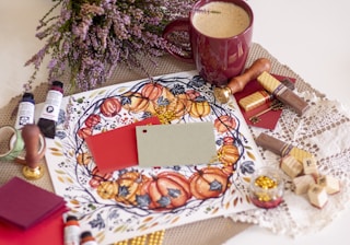 A cozy and artistic arrangement featuring a warm color palette. There is a decorative circular pattern with pumpkins on a piece of paper, alongside paint tubes, sealing wax, and a stamp. A cup of frothy coffee sits nearby, complementing the autumnal theme. Various crafting materials, including colored cards, gold beads, and stamps, suggest a creative environment. The scene is enhanced by dried flowers and textured fabrics.