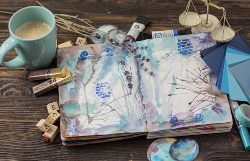 An open sketchbook with watercolor art and text lies on a wooden surface. The book is surrounded by a light blue coffee mug filled with coffee, chocolates, wooden alphabet stamps, a paintbrush, dried lavender flowers, a scale model, and various artistic tools like brushes, color samples, paint tubes, and round watercolor art pieces. The text in the sketchbook reads 'A baby is a tiny bit of heaven on earth.'
