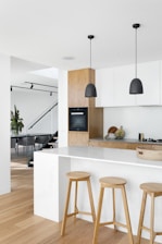A modern kitchen interior featuring a minimalist design. The space includes a large white marble island with three wooden stools. Two black pendant lights hang from the ceiling. The kitchen cabinetry is a combination of white and light wood. In the background, a staircase with glass railing leads to another part of the home.