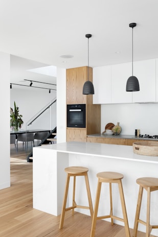 A modern kitchen interior featuring a minimalist design. The space includes a large white marble island with three wooden stools. Two black pendant lights hang from the ceiling. The kitchen cabinetry is a combination of white and light wood. In the background, a staircase with glass railing leads to another part of the home.