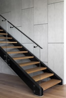 A modern staircase with wooden steps and glass side panels, set against a textured gray wall. The stairs feature a minimalist design with a sleek black railing and are integrated into a contemporary interior space.